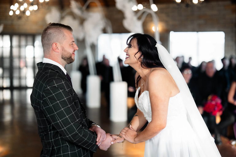 A bride and groom holding hands and facing each other during their ceremony, with guests behind them.