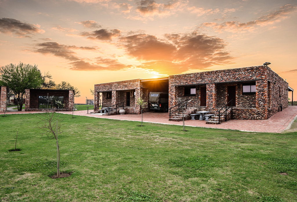 Brick accommodation with a covered stoep across a mown lawn at sunset.