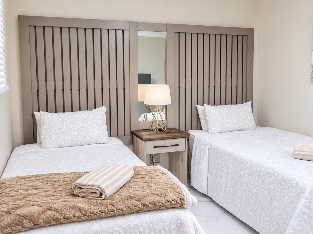 Two single beds in a Family Room bedroom, with a slatted headboard, a shared bedside table and lamp, and a rug underfoot.