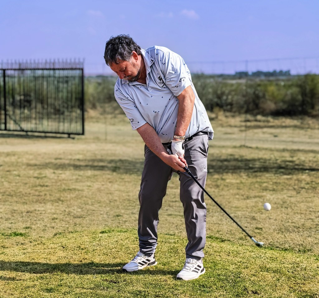 A golfer mid-swing on the grass at Goldfields Game Ranch, the ball in the air beside him.