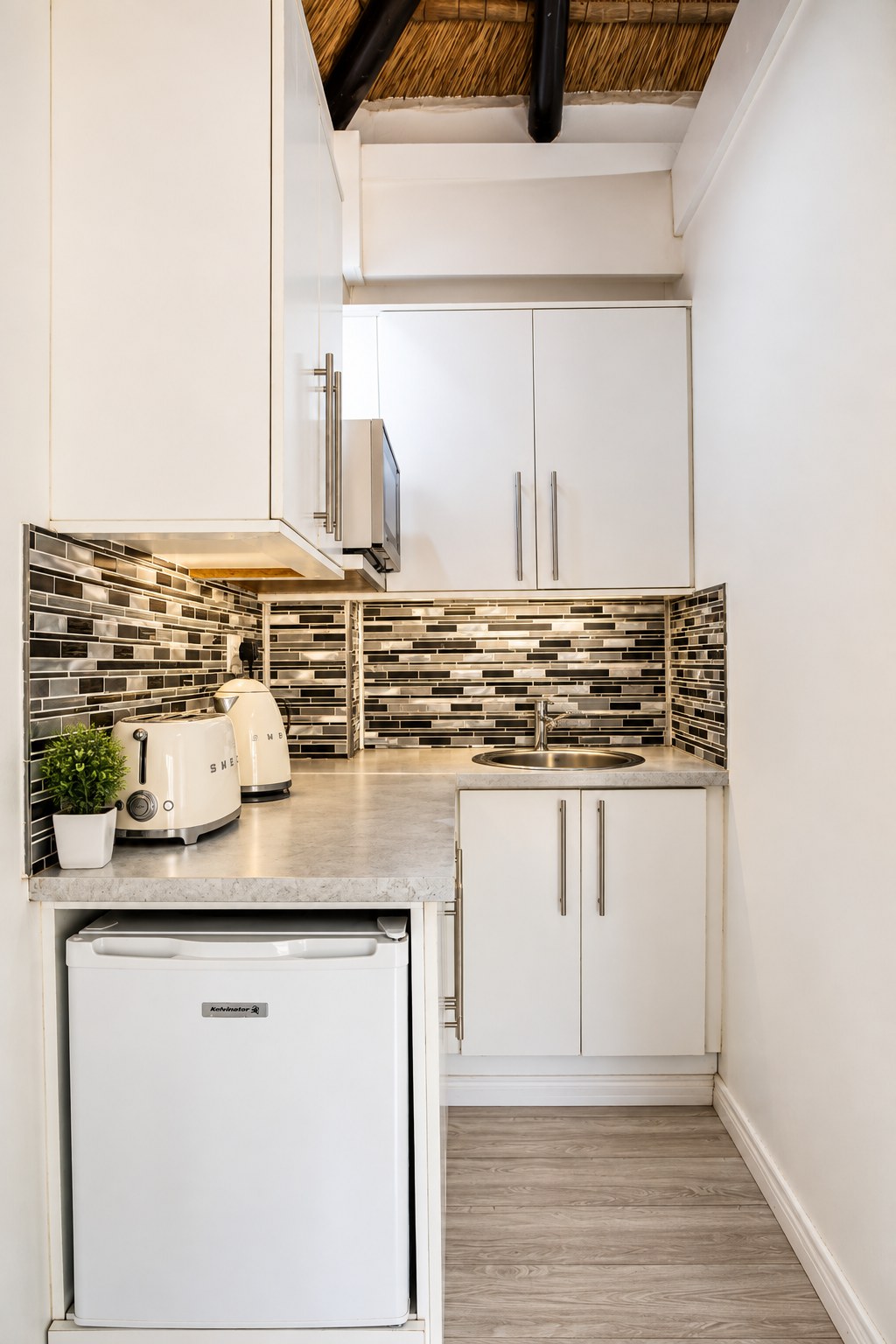 A Sandstone Room kitchenette with a bronze tiled splashback, microwave, kettle, toaster and bar fridge.