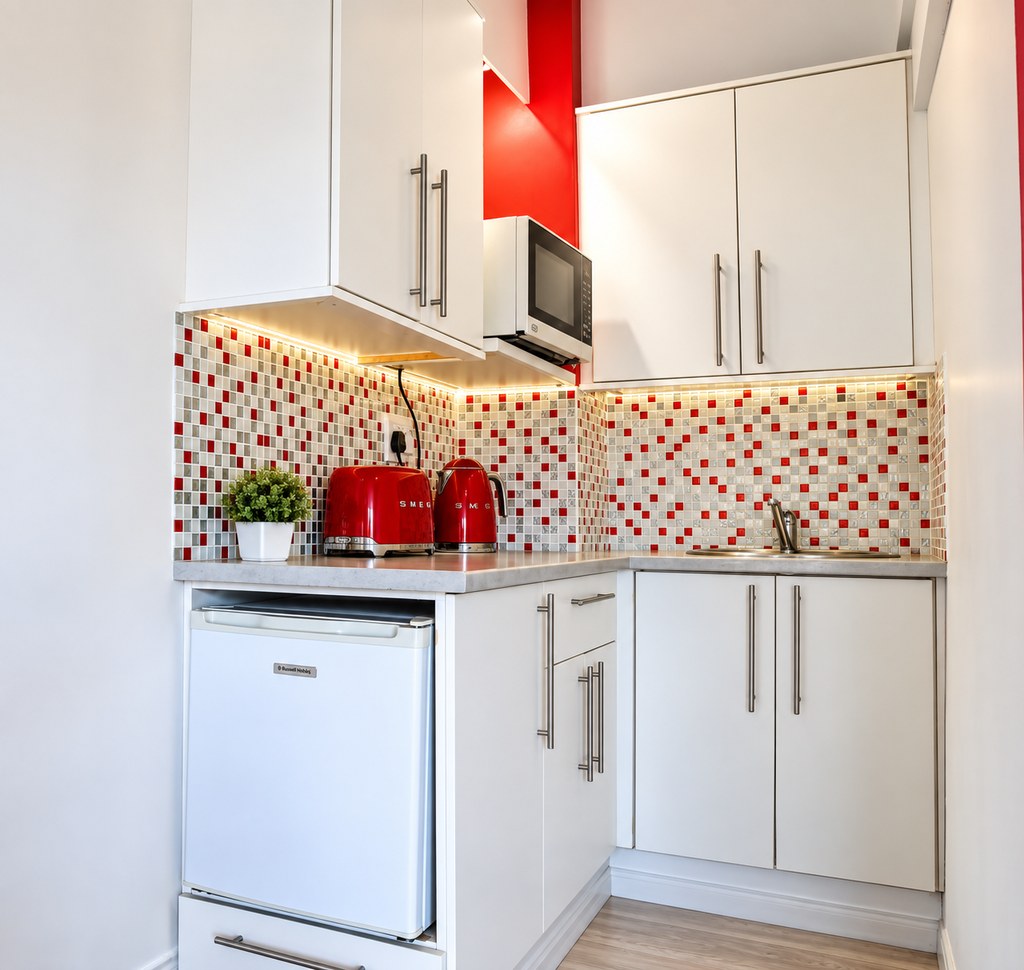 A Sandstone Room kitchenette with a red mosaic splashback, microwave, kettle, toaster and bar fridge.