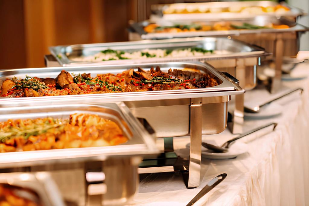 Chafing dishes of hot food set out along a draped buffet table, with serving spoons laid ready beside them.