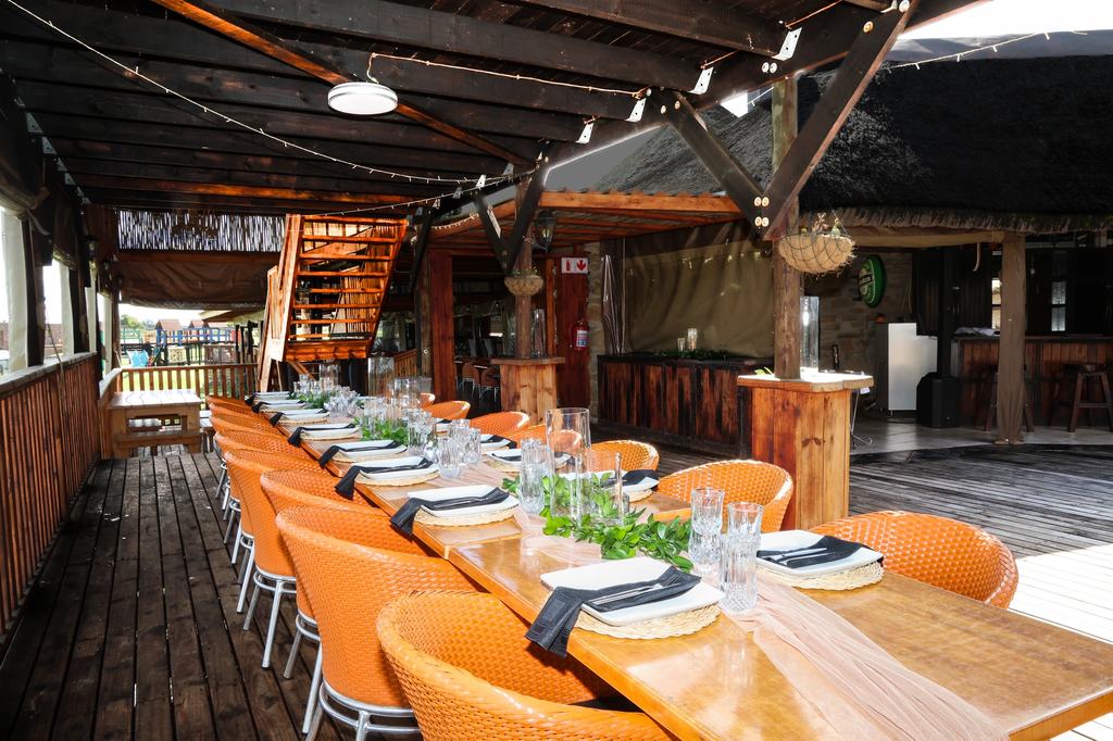 A long timber table laid for a function on the covered deck, set with woven placemats, folded napkins, glassware and a trailing greenery runner, with the bar behind it.