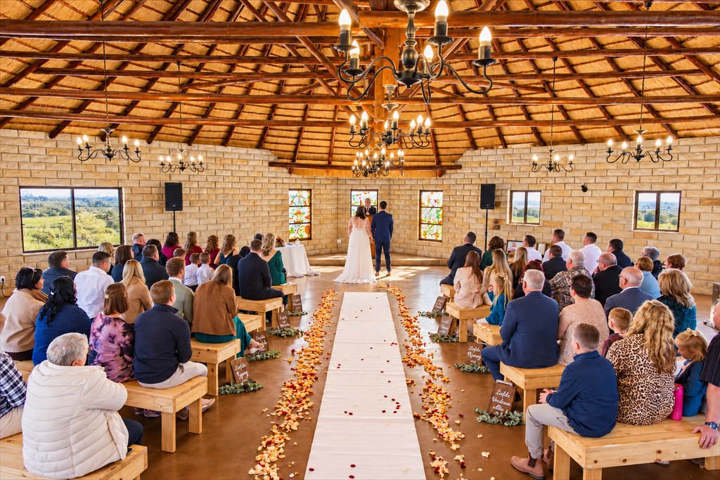 A wedding ceremony under the thatched pavilion roof, guests seated on timber benches.