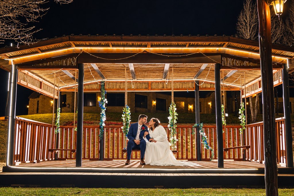 The lit timber gazebo in the evening, with the couple seated on the hanging swing.