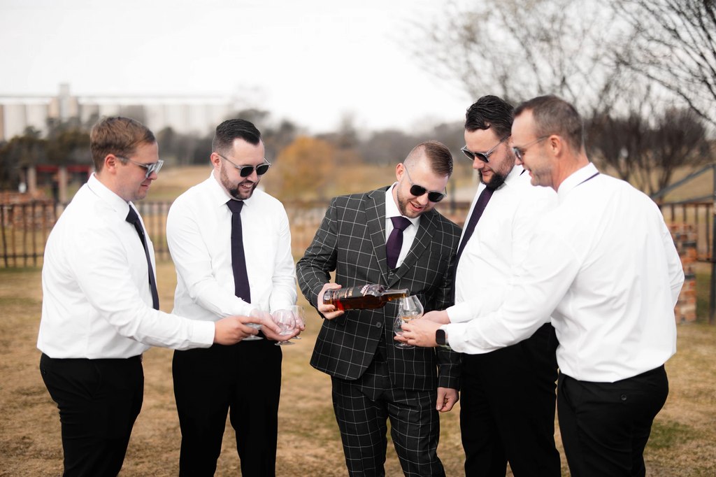 The groom and his groomsmen pouring a whisky together outdoors on the lawn.
