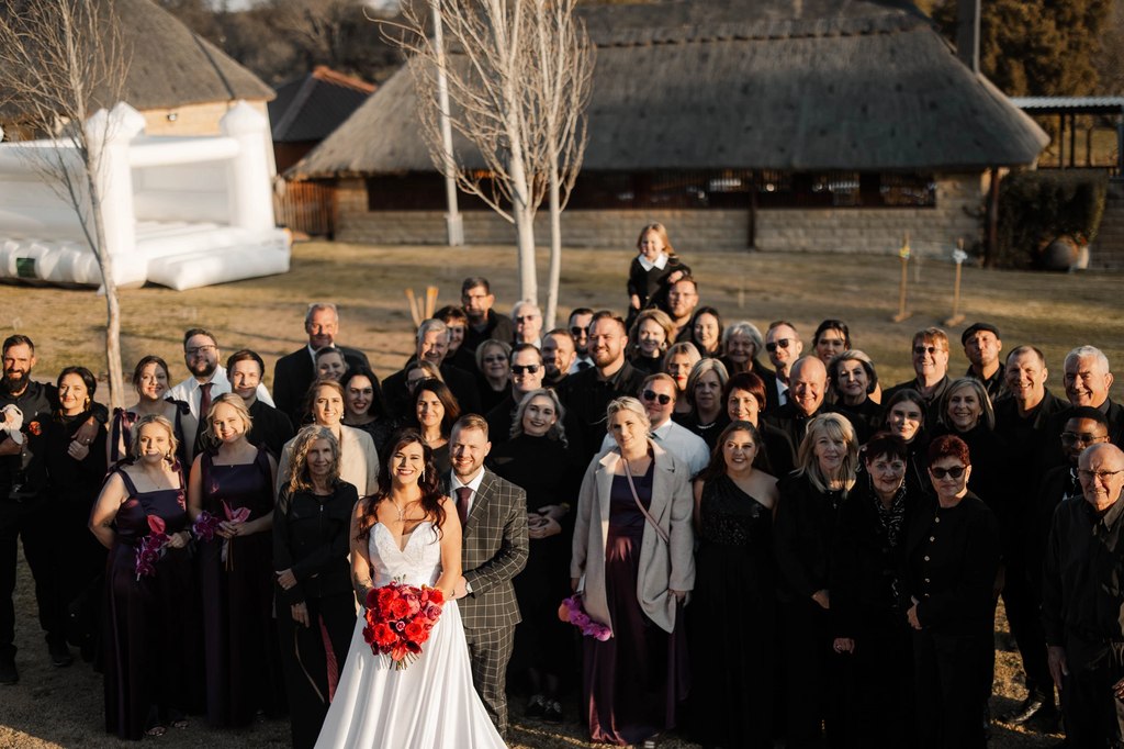An elevated group portrait of the whole wedding party and guests, with the thatched ranch buildings behind at Goldfields Game Ranch.