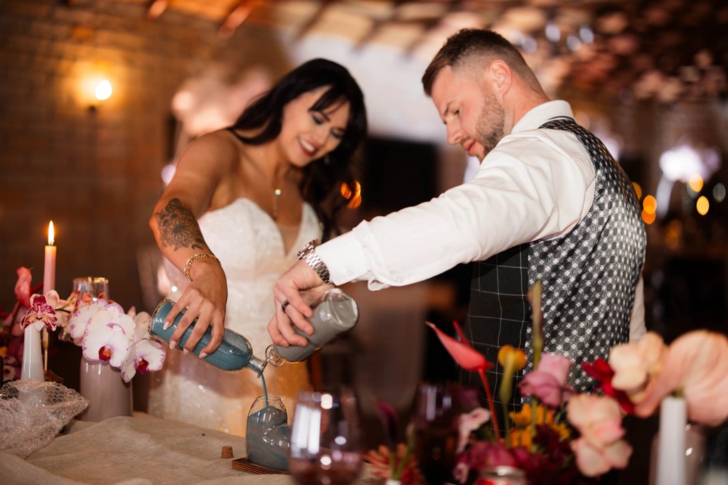 A bride and groom pouring a sand ceremony together at a decorated table.