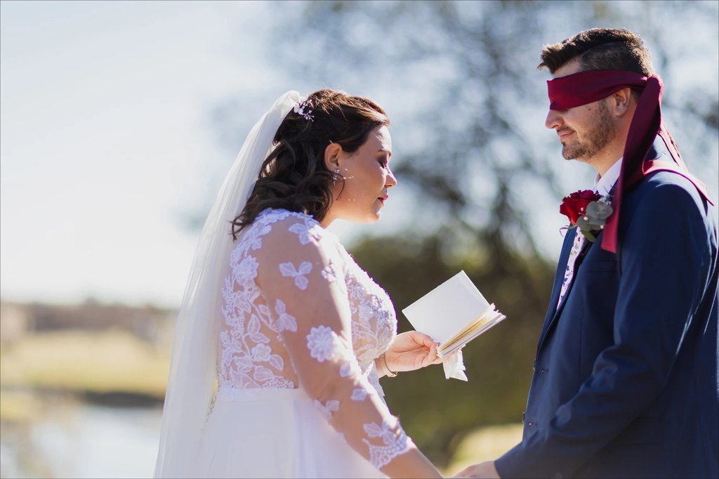 A blindfolded groom listening as the bride reads her vows beside the water at Goldfields Game Ranch.