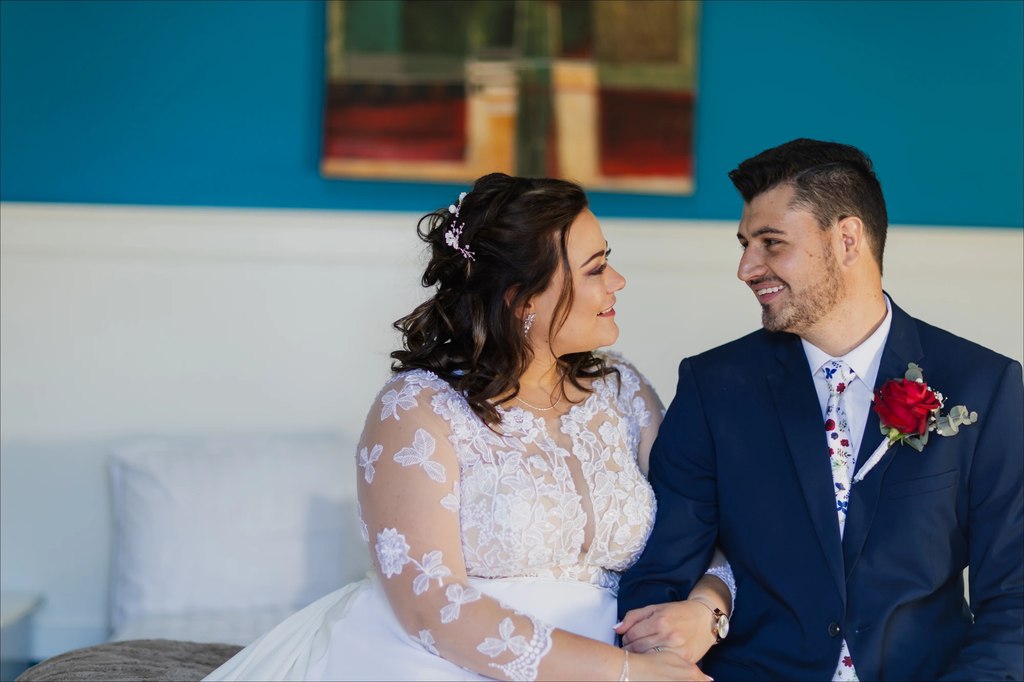 A bride and groom seated together indoors against a teal wall.