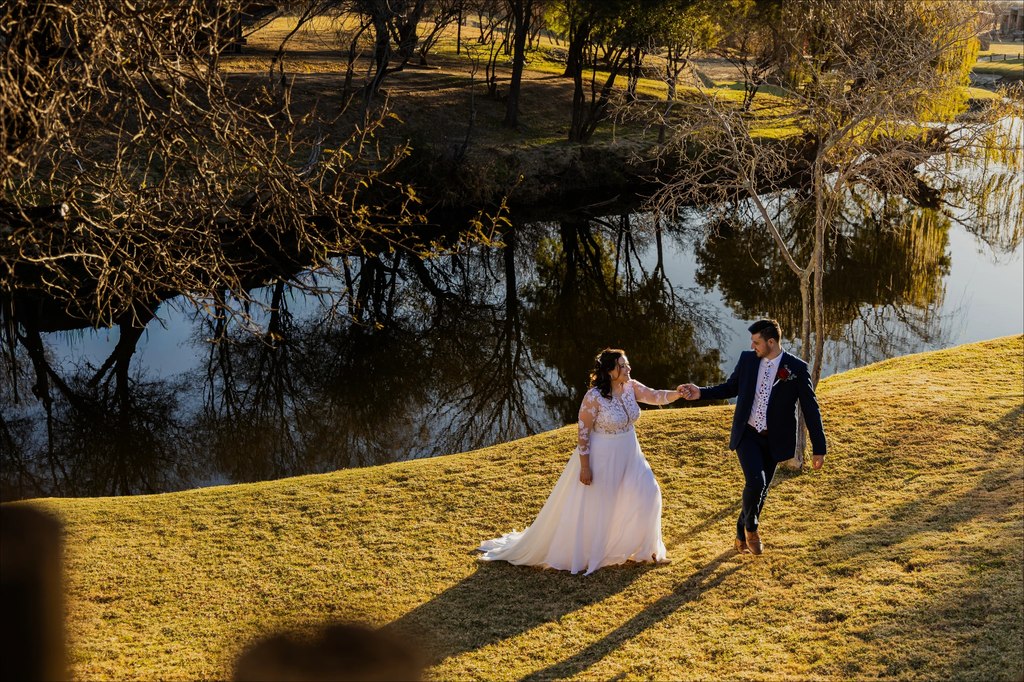 A bride and groom walking hand in hand along the grassed dam bank at golden hour at Goldfields Game Ranch.