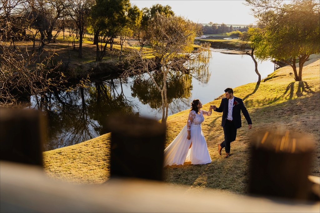 A bride and groom walking beside the dam at golden hour, seen past the deck railing at Goldfields Game Ranch.