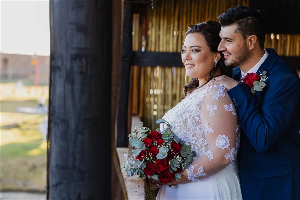 A bride holding a red-rose and eucalyptus bouquet beside the groom at the thatched lapa window at Goldfields Game Ranch.