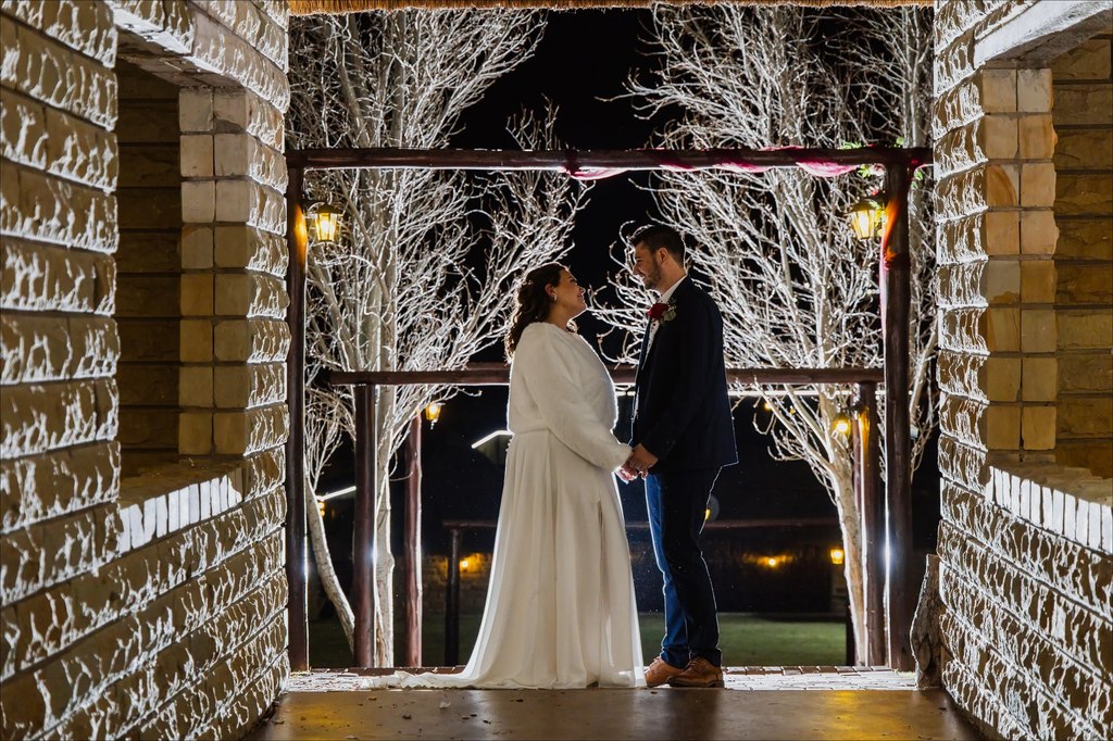 A bride and groom at night, framed by a sandstone arch with a lit white-branch arbour beyond at Goldfields Game Ranch.