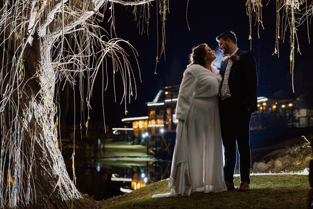 A bride and groom beneath an illuminated willow at night, the lit venue reflected in the water at Goldfields Game Ranch.