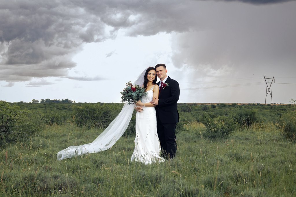 A bride and groom standing in grassland under a heavy storm sky, her veil trailing in the open Free State landscape.