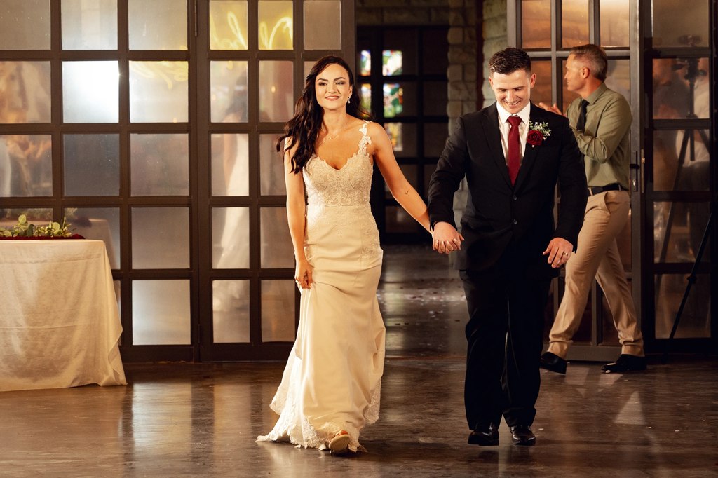 A bride and groom entering the reception hand in hand through the glazed doors, guests applauding.