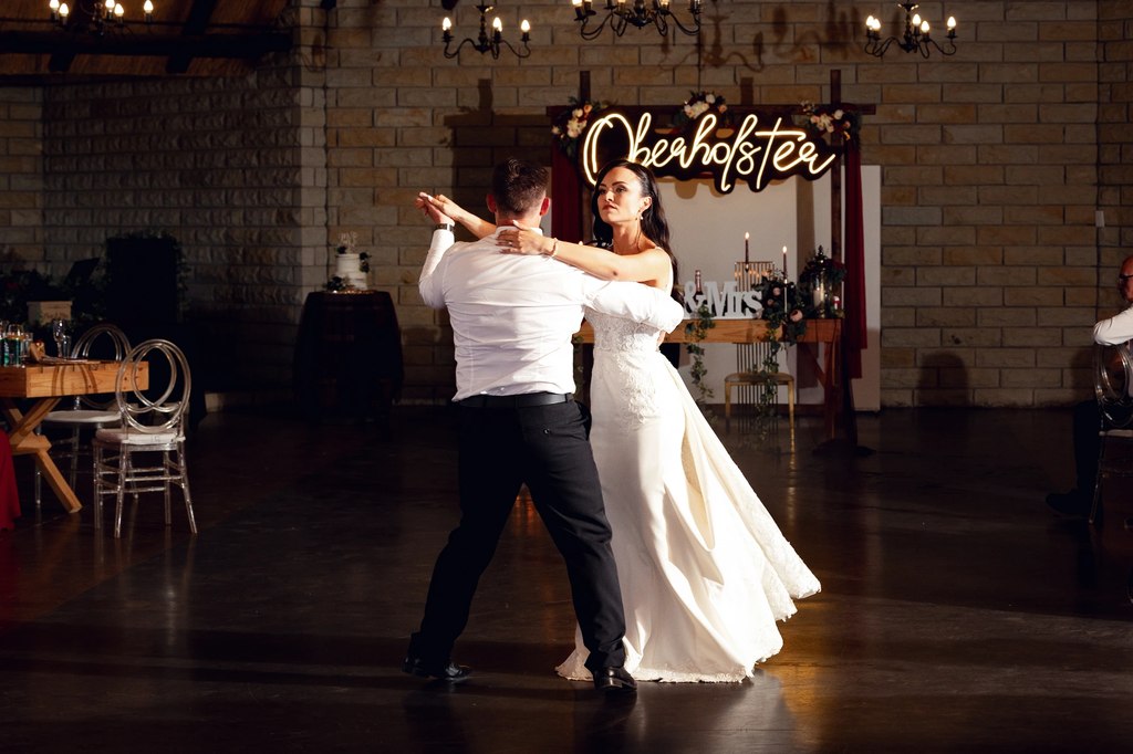 A bride and groom dancing on the hall floor beneath the illuminated reception backdrop.