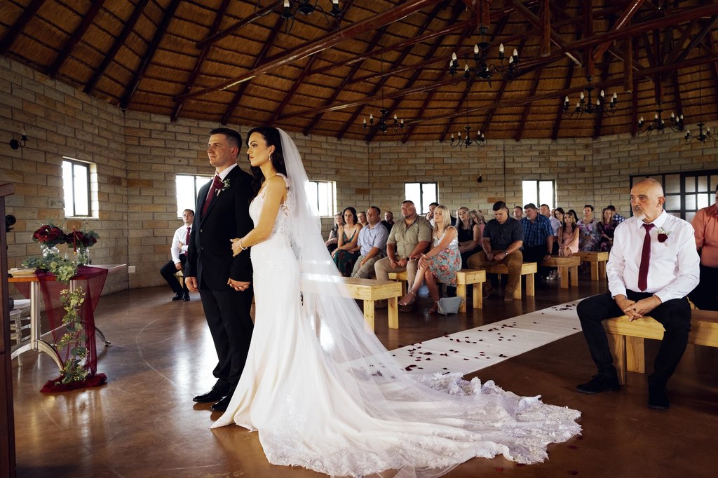 A bride and groom at the altar of the thatched round hall, her train spread behind her, guests seated on timber benches.