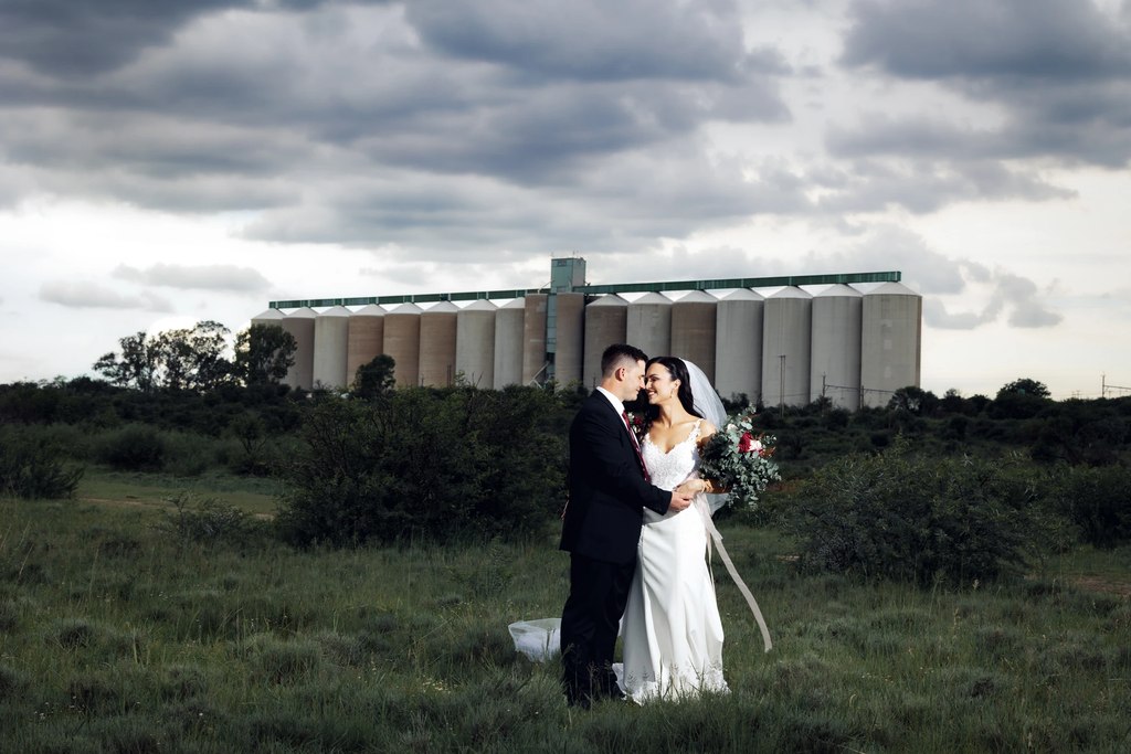 A bride and groom in grassland with grain silos behind them under a heavy storm sky in the open Free State landscape.