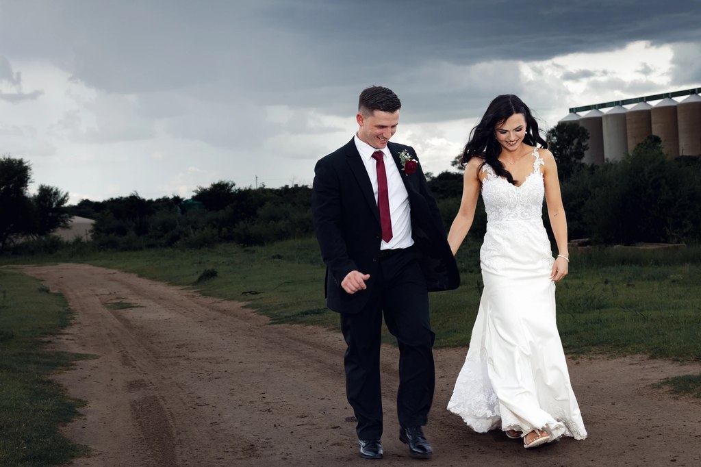 A bride and groom walking hand in hand down a farm track, grain silos behind them in the open Free State landscape.
