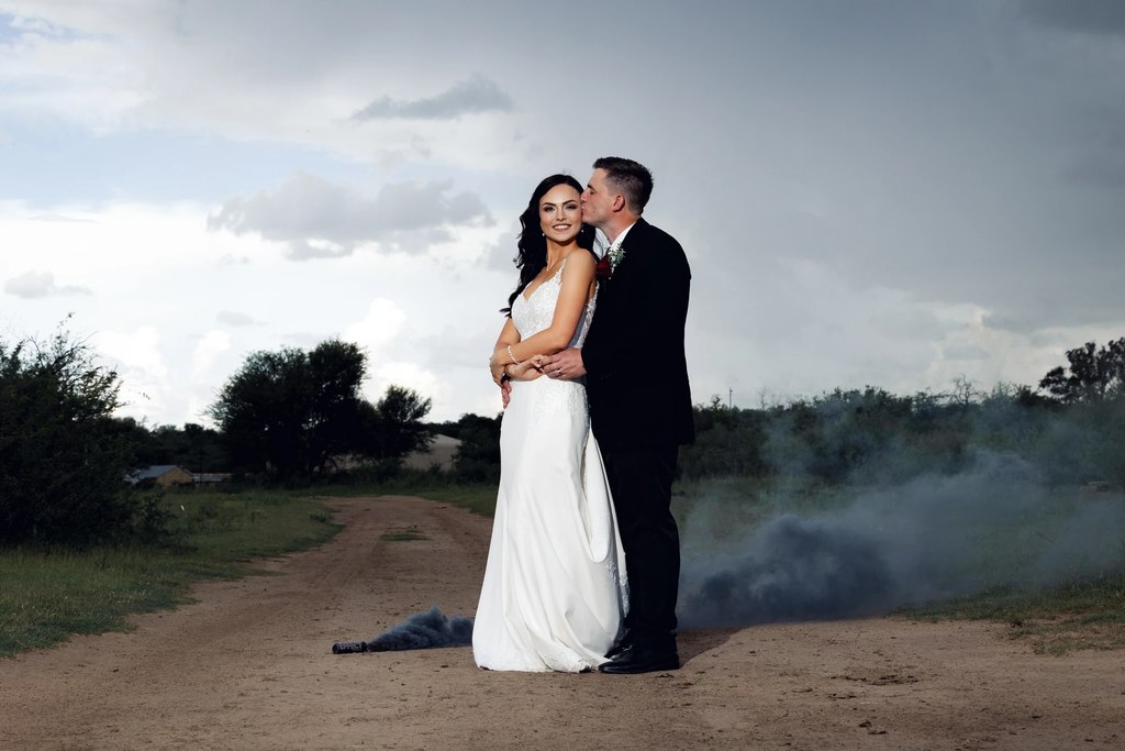 A bride and groom on a farm track with grey smoke drifting behind them in the open Free State landscape.