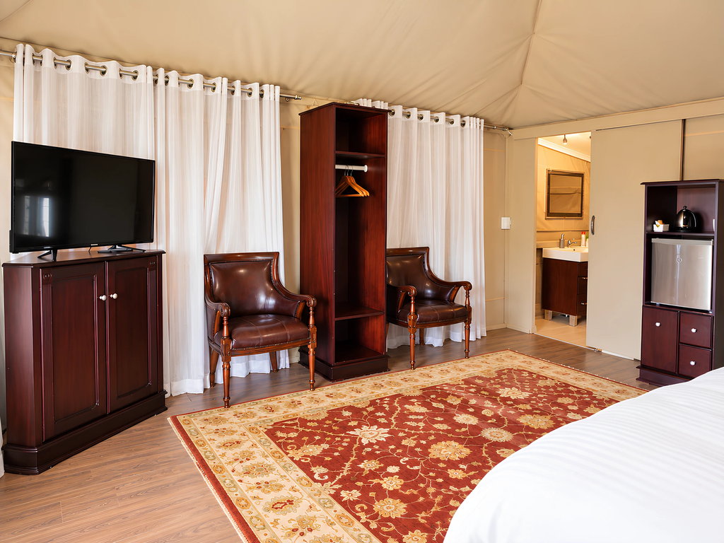 A flat-screen television, two armchairs and an open wardrobe inside a wooden house, with the bathroom beyond.