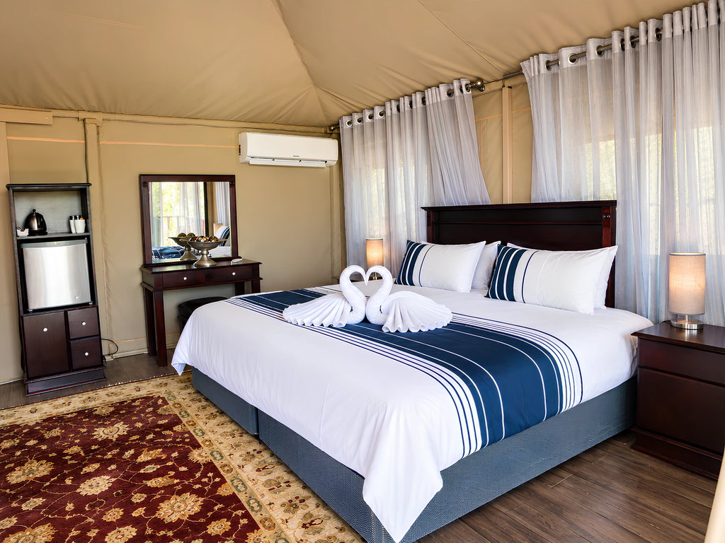 A made-up double bed inside a wooden house, with a dressing table, air conditioner and mini fridge.